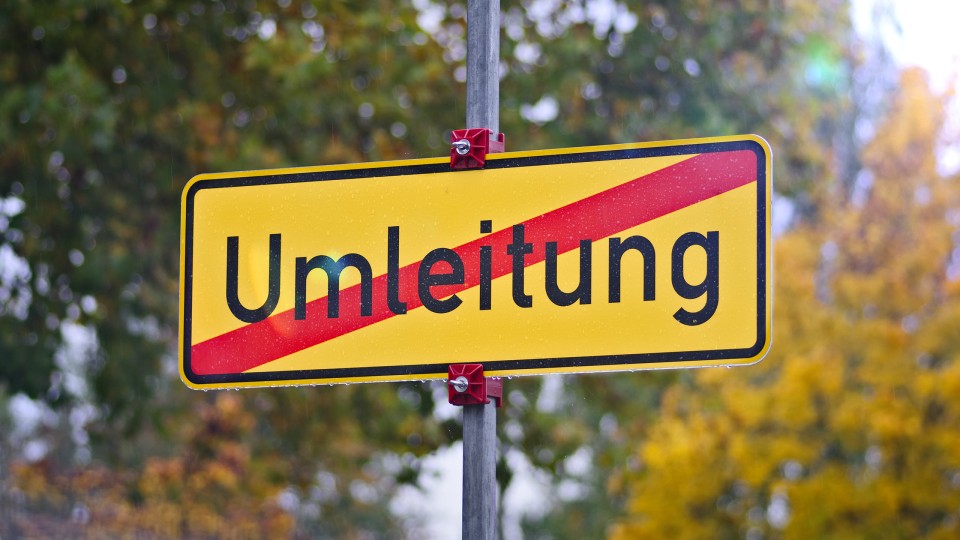 Baustellenschild an Straße bei grünem Baum im Hintergrund, Hinweis auf Busumleitung und Verkehrssperrung Erzhütter Straße.