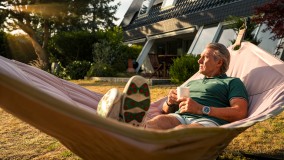 Gerry Ehrmann entspannt im Garten in einer Hängematte. Er hält eine Tasse in der Hand und wird von der Sonne angestrahlt.