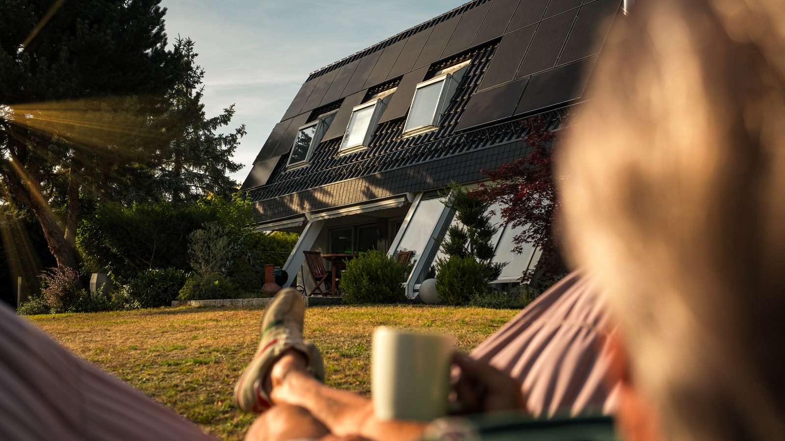 Person entspannt mit Tasse im Garten vor Einfamilienhaus mit moderner Photovoltaik-Dachanlage bei Sonnenuntergang.