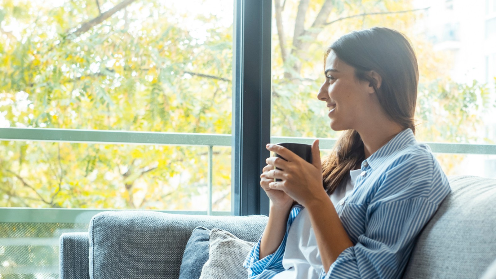 Eine junge Frau sitzt mit einer Tasse auf der Couch vor einer Fensterfront und schaut nach draußen.