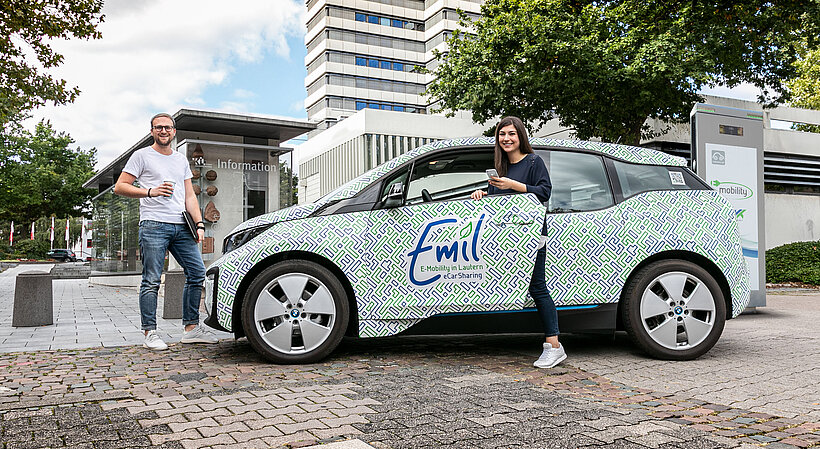 Personen an BMW i3 Emil eCarSharing von SWK, Frau hält Handy in Hand und steht in der Tür, Mann steht vor Auto und hält Tasse und Mappe in seiner Hand