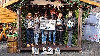 Personen stehen in einer weihnachtlich dekorierten Holhütte. Zwei halten einen Scheck in der Hand, die anderen gefüllte Beutel.