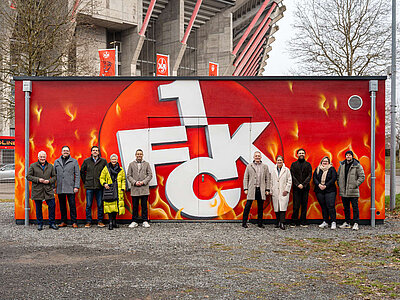 Personen stehen vor der Fernwärmezentrale am Stadion. Die Fassade ist mit einem Graffity besprüht, welches das FCK-Logo umgeben von Flammen zeigt.