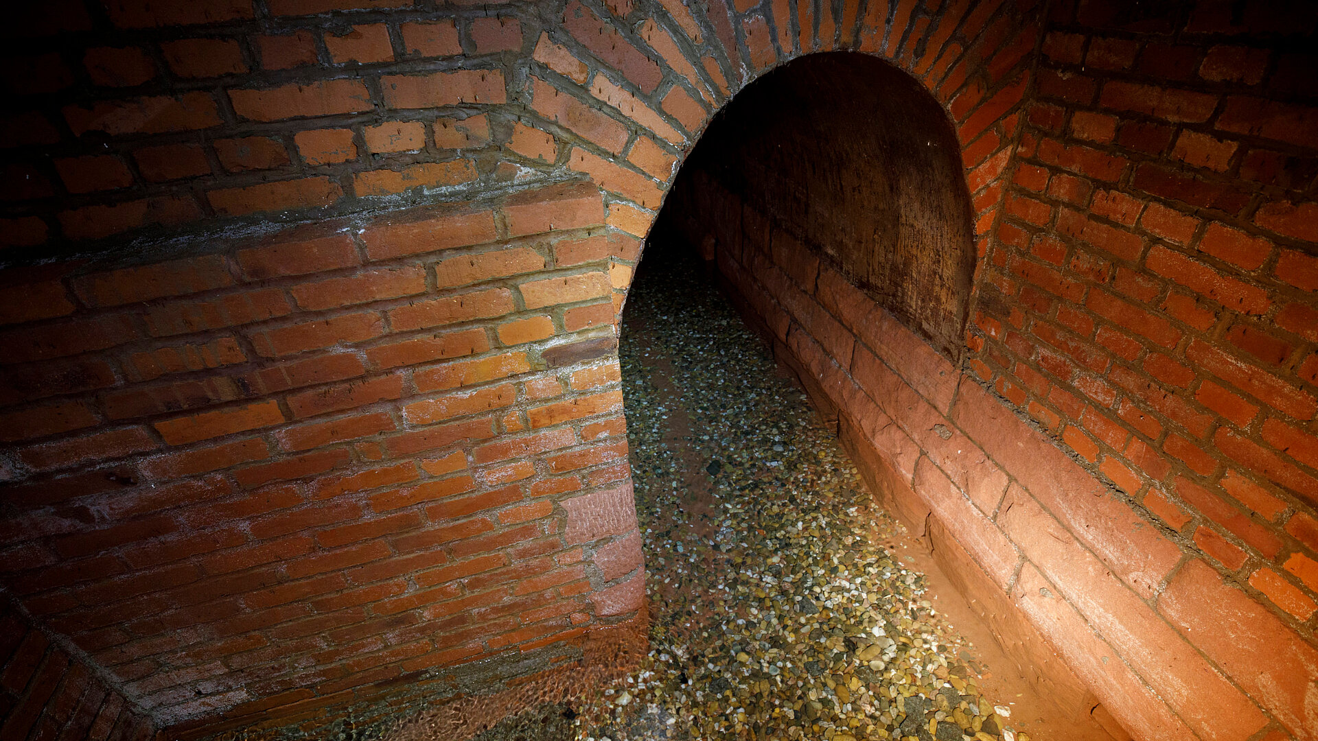 Gemauerter Gewölbetunnel mit flachem Wasserverlauf und Kiesbett.