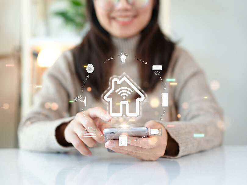 Frau sitzt mit Handy in der Hand am Tisch. Darüber liegt eine Illustration. Ein Haus kreisförmig umgeben von verschiedenen Symbolen im Haushalt.