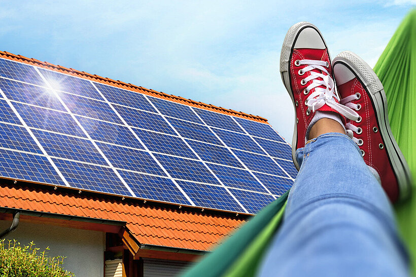 Beine und Füße einer Person auf Hängematte mit Blick auf Haus mit Solaranlage