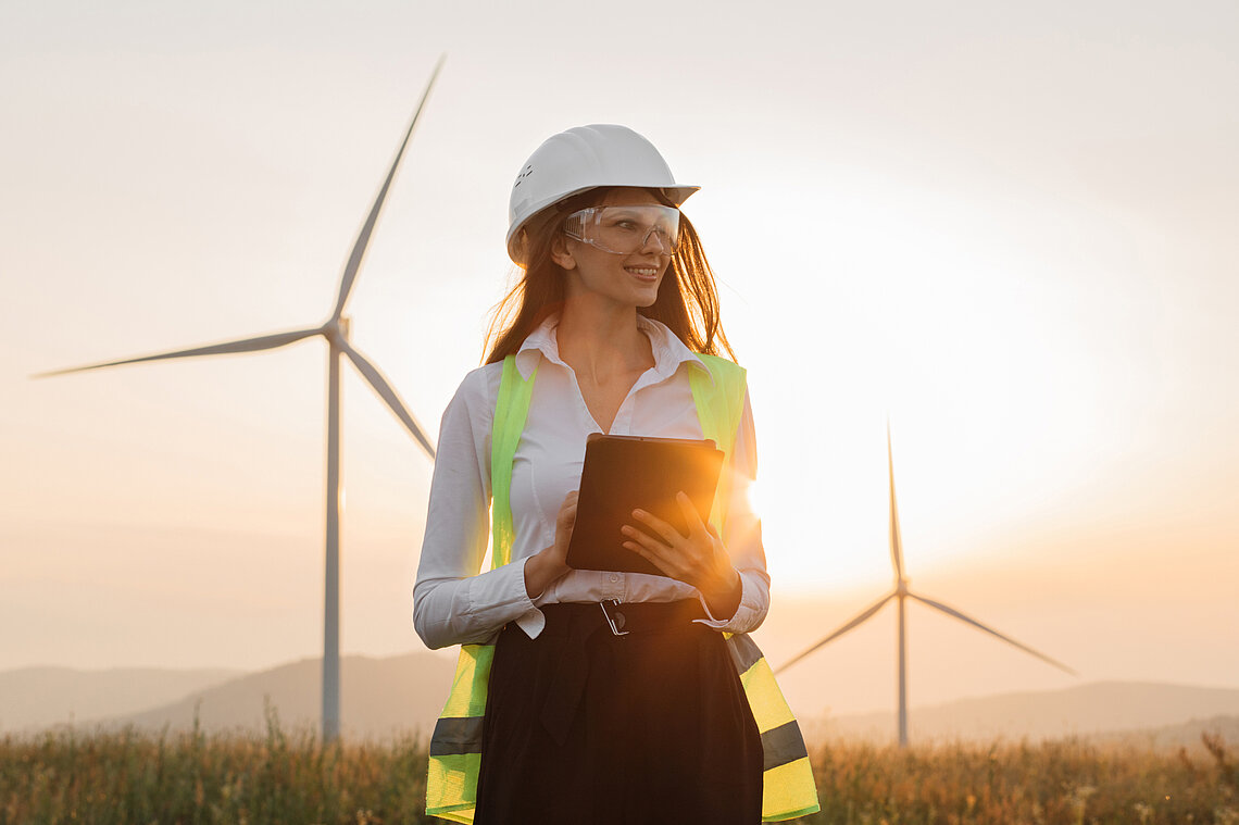Frau vor Windrädern in Schutzweste und Schutzhelm- und Brille mit Tablet in ihrer Hand