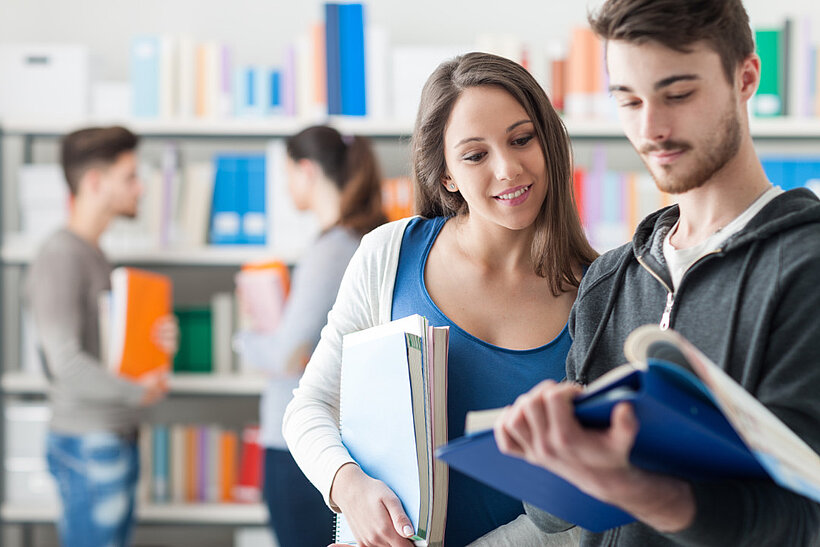 Personen in Bücherei, Mann zeigt Frau etwas in Buch und Frau schaut in sein Buch und hält weitere Bücher unter ihrem Arm