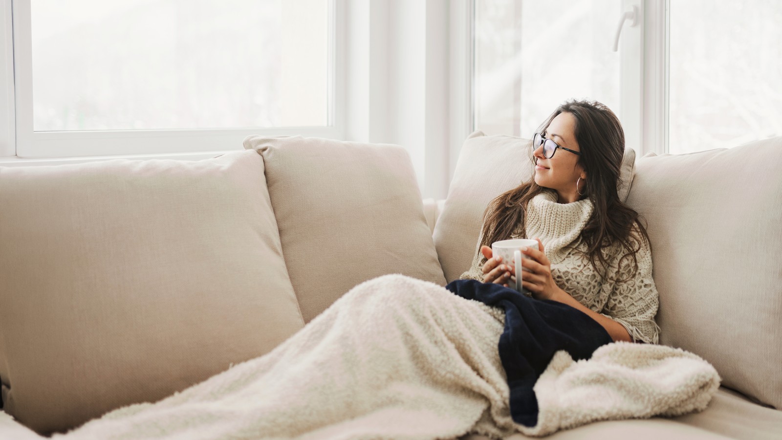 Frau entspannt mit Decke über ihrem Schoß und einer Tasse in der Hand auf der Couch. Sie schaut glücklich aus dem Fenster.